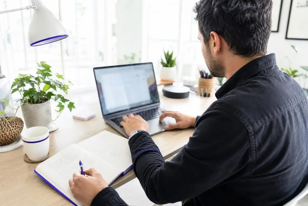 A man is working on a laptop and taking notes in a notebook.