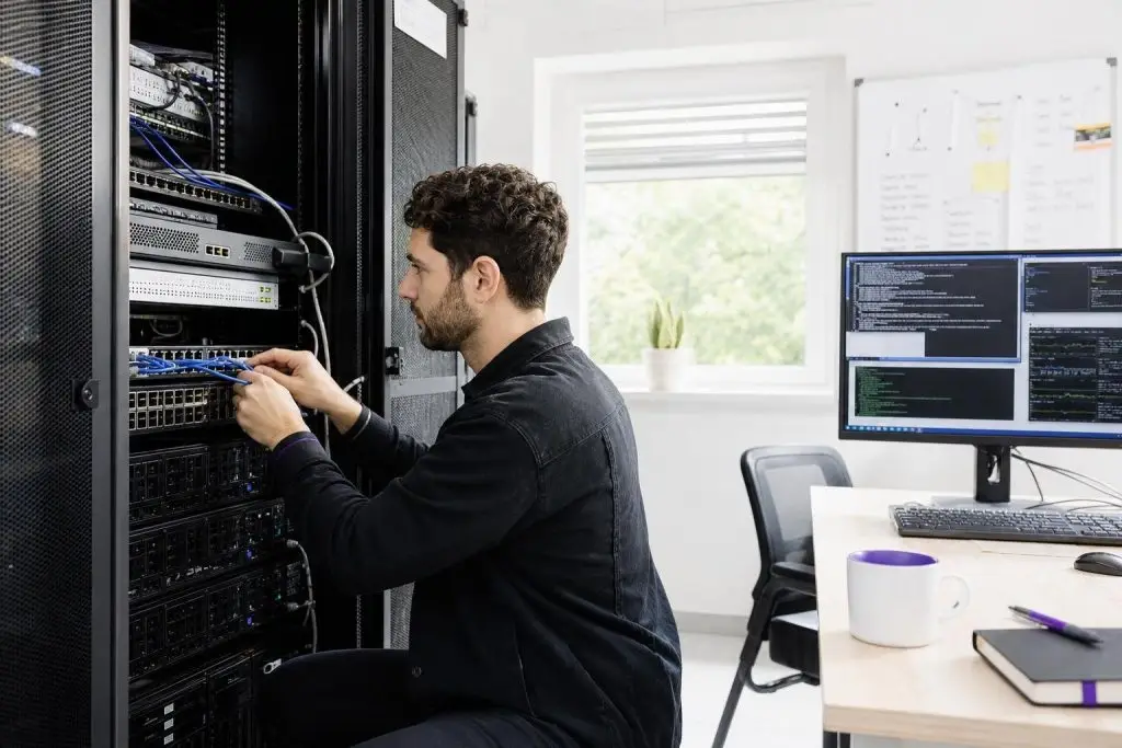 A man is working on a server rack in an office with a computer screen.