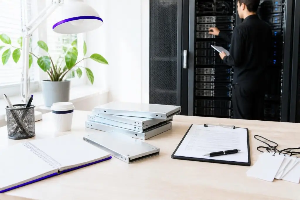 A man works on a server rack in a bright office with a desk and note-taking materials.