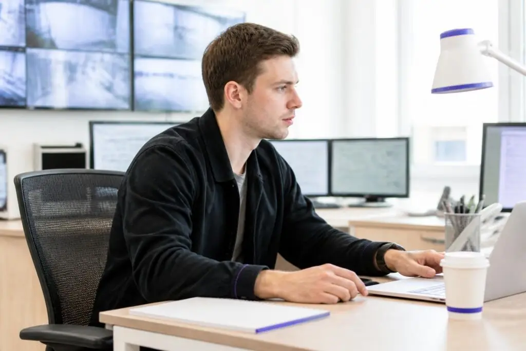 A man working on a laptop in an office with multiple screens.