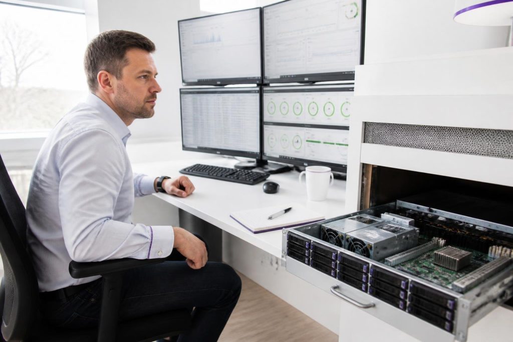 A man sits at a desk with multiple monitors and an open server chassis.
