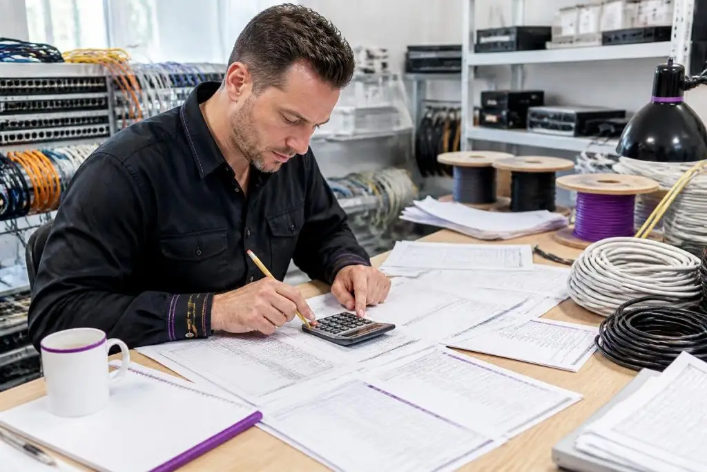 A man calculates something with a calculator at a table full of papers and cables.