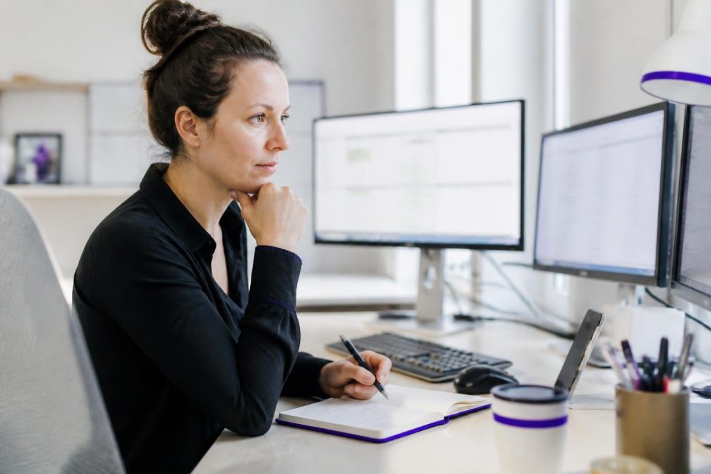 A woman sits at a desk with multiple monitors, taking notes.