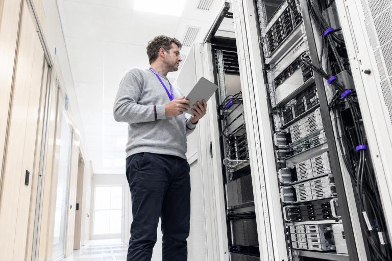 A man with a tablet stands in front of server racks in a data center.