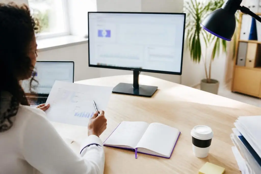A person is looking at a document with charts at a desk with a monitor and laptop.